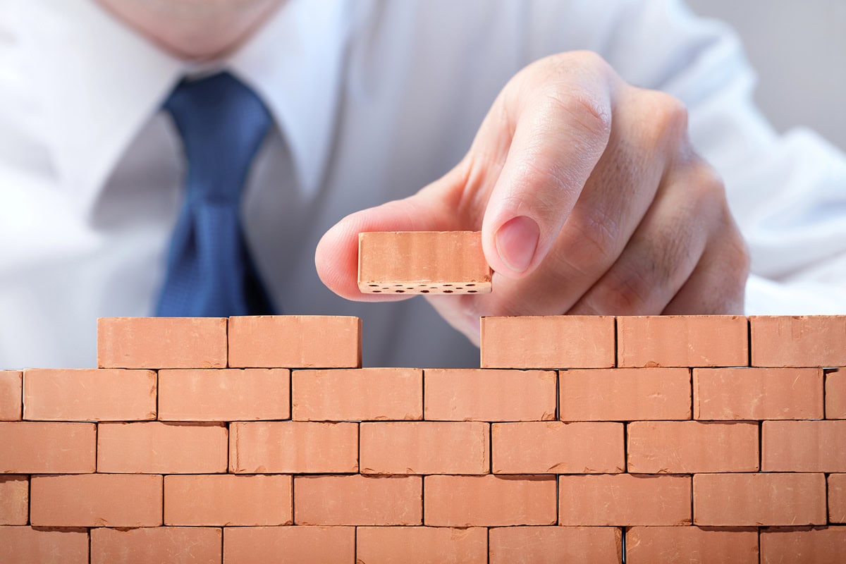 a businessman places a tiny brick into a tiny brick wall.