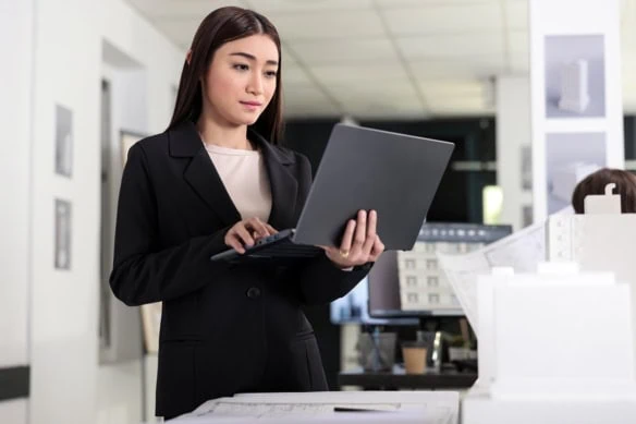 A young architect holds her laptop while standing.
