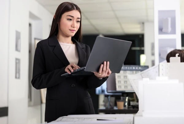 A young architect holds her laptop while standing.