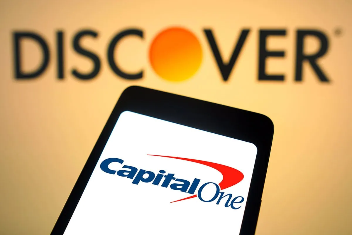 7 Best Value Stocks to Buy Right Now 3 the discover logo is shown above a phone displaying the capital one logo representing a merger between the two companies.