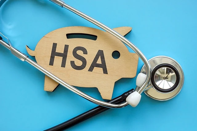 HSA Contribution Limits for 2026 + 2027 2 a wooden cutout of a piggy bank that says H S A sits against a blue background with a stethoscope.