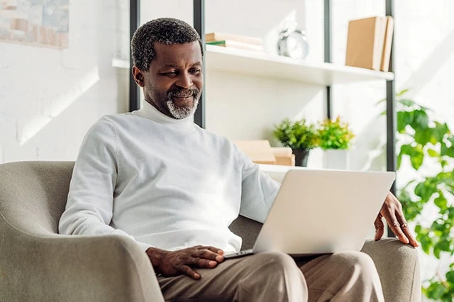 a black man wearing a white sweater smiles while looking at his laptop.