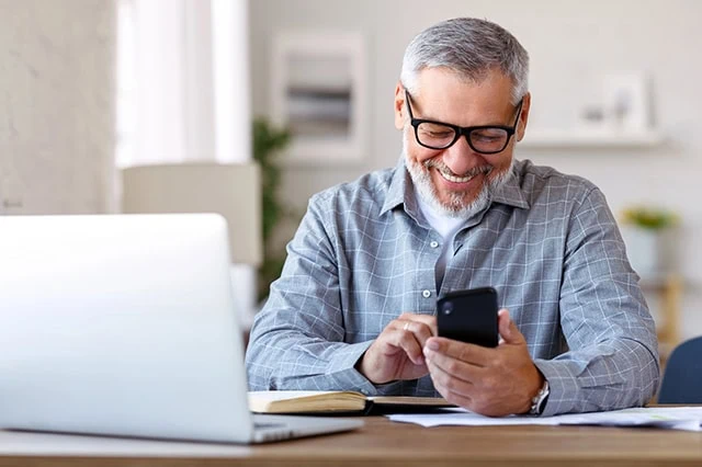 5 Social Security Moves Every High-Net-Worth Individual Should Know 4 a man smiles while looking down at his smartphone at his desk at home.