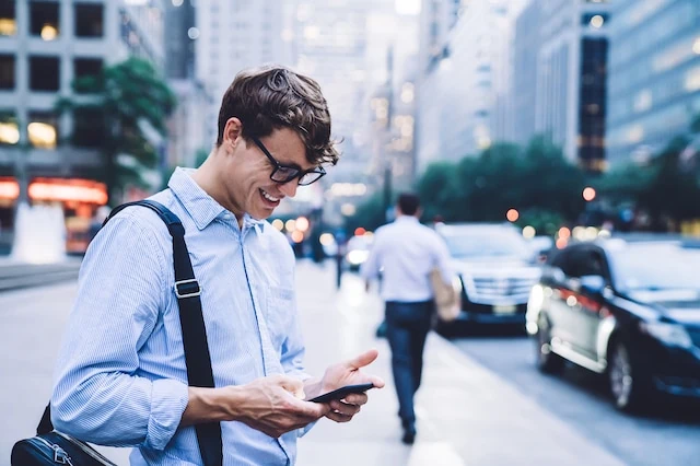 Young contemporary male worker in shirt and eyeglasses messaging on phone standing against high buildings of New York looking happy